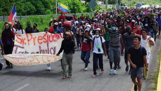 Imagen que muestra a un grupo numeroso de personas migrantes que forman una caravana, no se perciben sus rostros, pero los que van hasta adelante llevan una pancarta con un mensaje que no se aprecia porque está doblada, y se ven algunas banderas de sus países de origen.