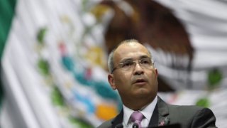 Fotografía de un hombre maduro, de lentes y con traje frente a una bandera de México