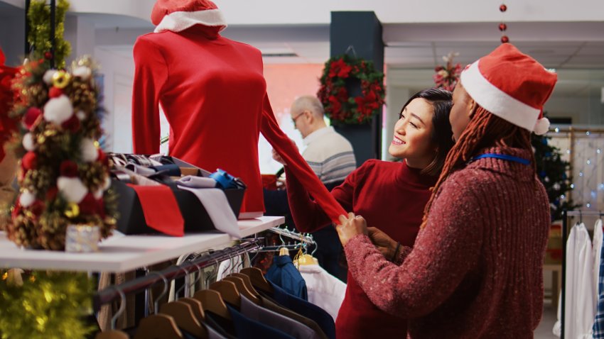 Friendly retail assistant in festive ornate shopping mall fashion shop showing client beautiful red garments, ready to be worn at Xmas themed holiday events during winter season