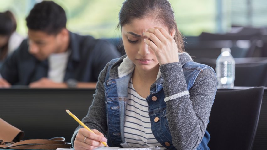 A young woman sits at a table in her college lecture hall and looks down in concentration as she takes a difficult exam.  She frowns and rests her head on her fingertips.