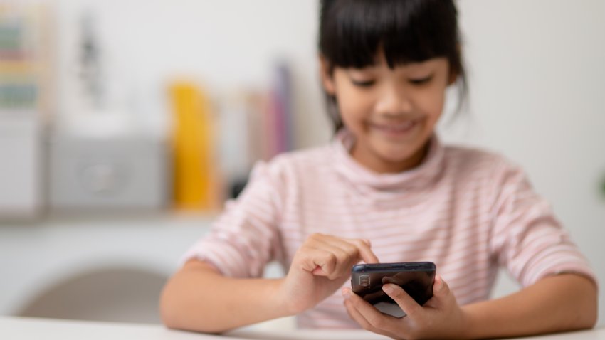 Cute little girl uses smartphone while sitting at the sofa in the living room. Child surfing the internet on mobile phone, browses through internet and watches cartoons online at home