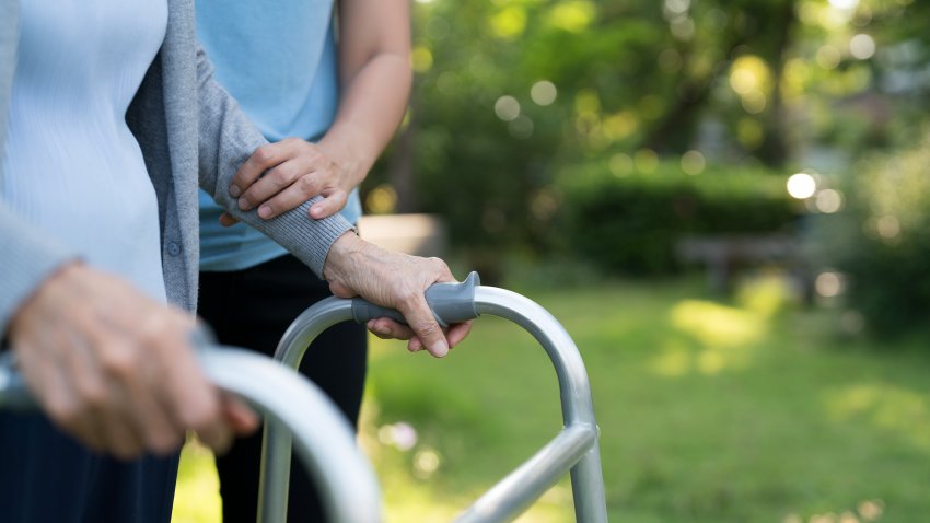 Image of an unrecognizable senior woman being assisted to walk using a walker by caregiver at park.