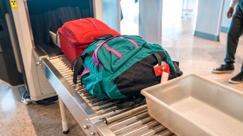 A colorful backpack and a small suitcase coming out on a roller conveyor after being scanned in a x-ray machine for security. A plastic tray in front. 3/4 length shot.