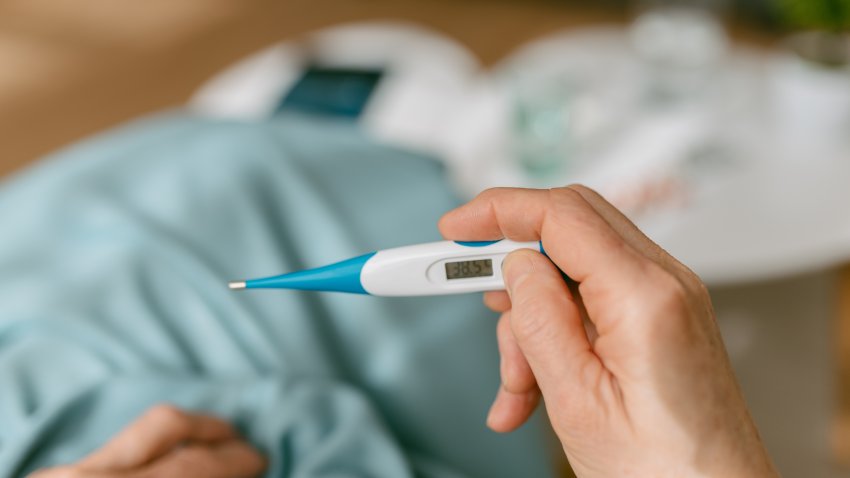 Over-the-shoulder view of an unrecognizable woman looking at the thermometer because she is sick while sitting on the sofa covered with a blanket.