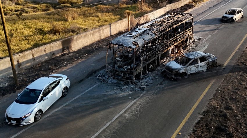 This aerial view shows a car passing by a burned bus and car, allegedly set on fire by organised crime groups in response to an operation to arrest a high-priority security target, on a highway near Acatlan de Juarez, Jalisco state, Mexico on February 22, 2026. The Mexican army announced that it had killed powerful drug lord Nemesio "El Mencho" Oseguera in an operation that sparked a wave of violence in various parts of the country on February 22, 2026. Gunmen retaliating for the raid blocked more than 20 roads in western Jalisco state, which includes Tapalpa, with burning cars and trucks. The violence spread to other states as well. (Photo by Ulises RUIZ / AFP via Getty Images)