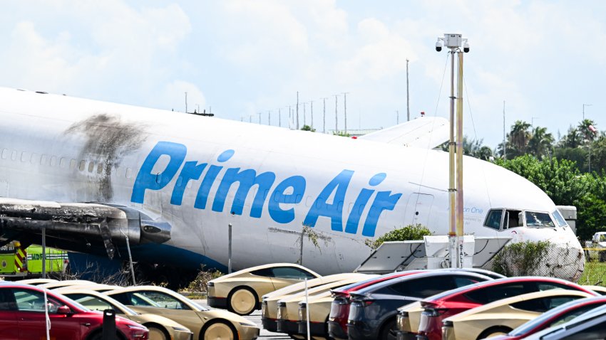 A parking lot filled with Tesla Cybercabs is seen in the foreground as the nose of a Prime Air cargo plane rests on the ground after the aircraft overran the runway and struck cars the previous day at Miami International Airport in Miami, Florida, on September 7, 2026. At least five people were killed when an Amazon cargo plane overshot a runway after it landed September 6 at Miami International Airport, crashed into vehicles and caught fire, officials said. Miami-Dade County fire chief Ray Jadallah told reporters that in addition to five fatalities, three people were transported to trauma centers "in critical condition" and two more were treated at a local hospital. "The airplane did strike a couple of vehicles," which left "a couple of victims that were actually trapped in the vehicles," Jadallah said. (Photo by CHANDAN KHANNA / AFP via Getty Images)